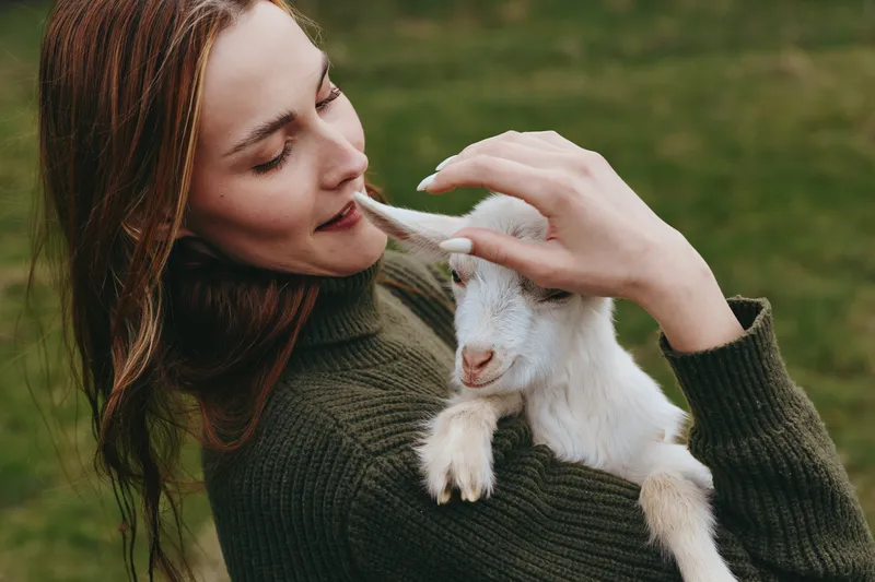 woman outdoors holding goat kid in gentle embrace, warm sweater, sunlit grass, tender interaction, natural colors, calm atmosphere, authentic lifestyle moment, close bond between human and animal,