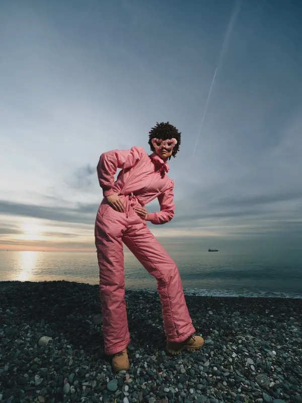 Person in pink snowsuit posing on a rocky beach by the sea during sunset, outdoor fashion moment with rugged shoreline and dramatic sky, lifestyle and adventure vibe