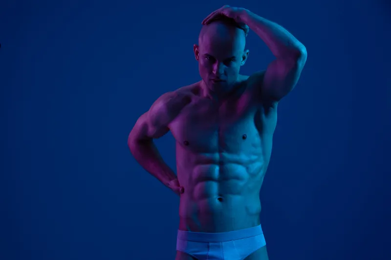 Athletic man posing dramatically against a dark blue background, showcasing strong physique and expression. The image features vibrant contrasting lights.