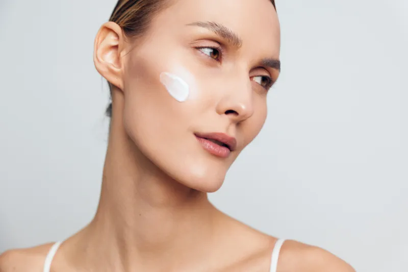 Young woman with clear skin applying face cream on cheek, natural beauty concept, soft lighting, minimal makeup, smooth complexion, skincare routine, neutral background
