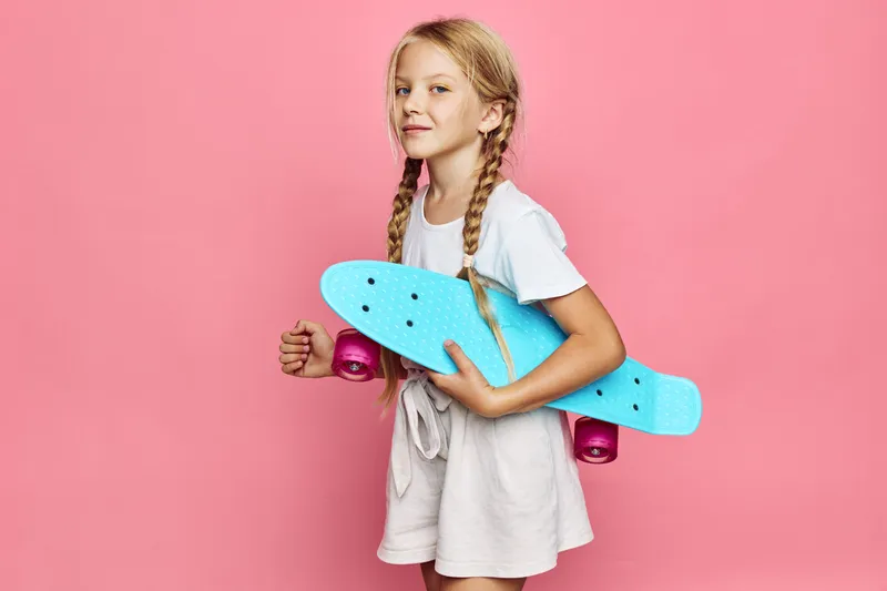 Young girl with braided hair holds blue skateboard and smiles while standing against pink background. Casual outfit with confident expression. Indoor studio shot.