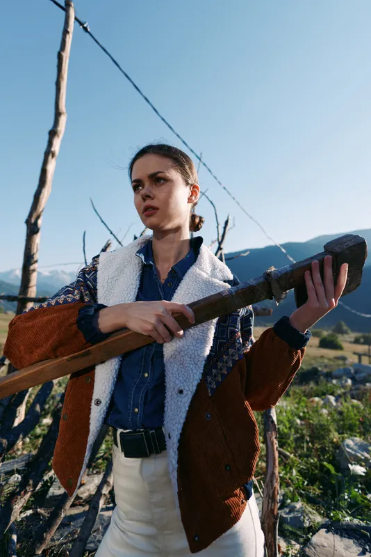 Woman holding a pole outdoors in mountains, rural portrait of a young female worker in jacket and denim, standing in a field with determined expression under clear sky and sunlight
