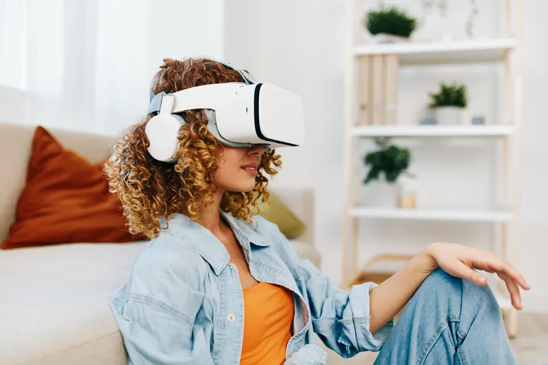 Улыбающаяся женщина наслаждается играми в виртуальной реальности дома в очках VR на футуристическом диване