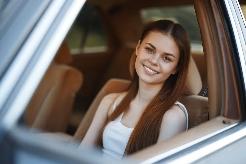 Smiling young woman sitting in a car, wearing a casual white tank top, showcasing natural beauty and joy in a sunlit environment.