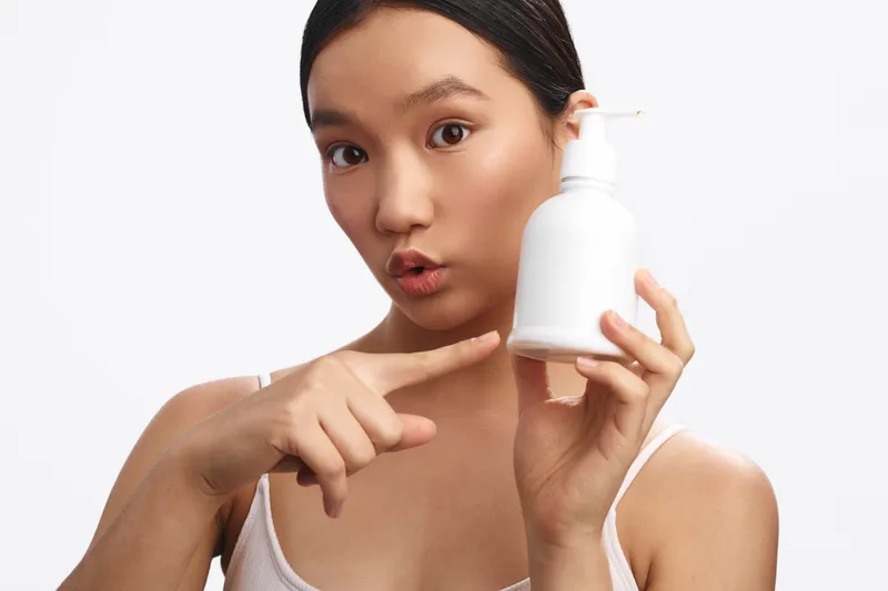 asian woman uses skincare product in a bright studio, holding bottle and pointing with a curious expression, promoting healthy skin and personal care in a clean modern setting.