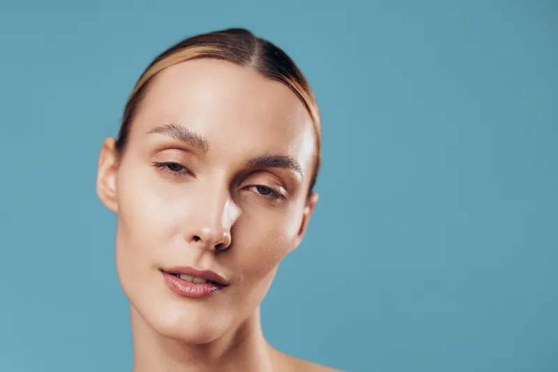 Close-up portrait of a young woman with slicked-back hair and natural makeup posing against a plain blue background with a calm and serene expression.