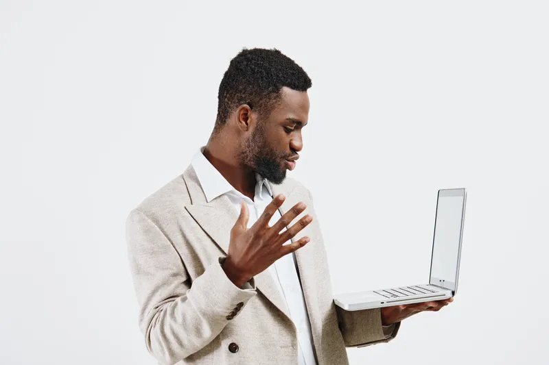 Businessman with dark skin holding a laptop and looking at the screen, dressed in beige blazer and white shirt, standing against plain white background. Modern workspace concept