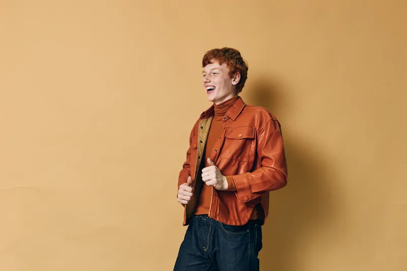 Young man with red hair laughing in casual orange jacket and black pants standing against beige background. Cheerful male model in stylish outfit posing with joyful expression.