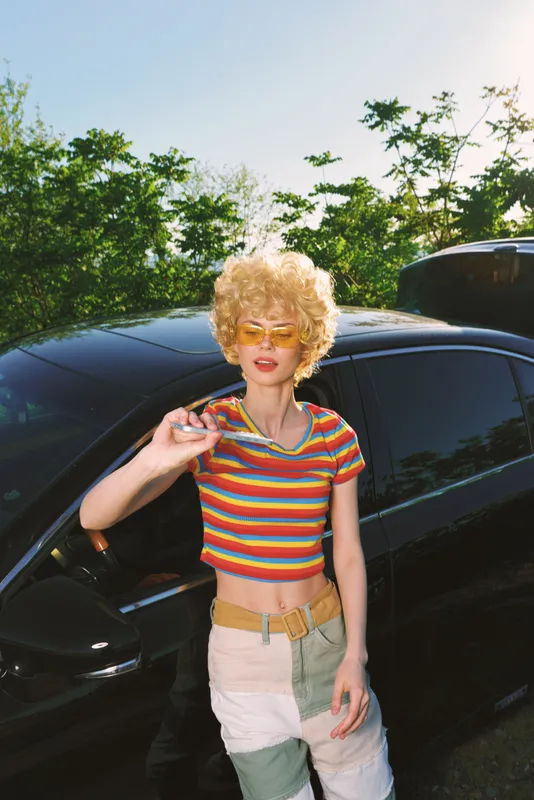 Woman outdoors, retro fashion vibe with striped crop top, blonde curly hair, yellow glasses, leaning by a glossy car, sunny day, vibrant colors, casual lifestyle moment, carefree mood