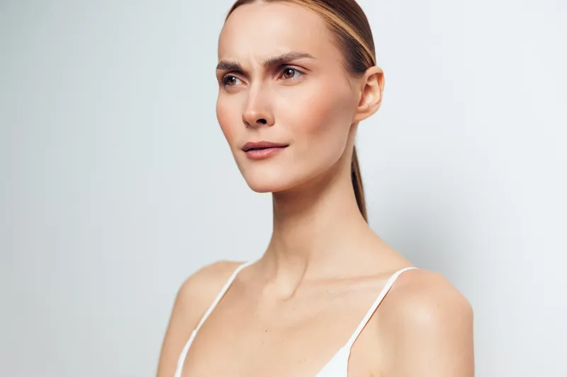 A focused young woman with a sleek ponytail gazes to the side, wearing a white tank top in a clean studio, conveying concentration and calm during a skincare study.