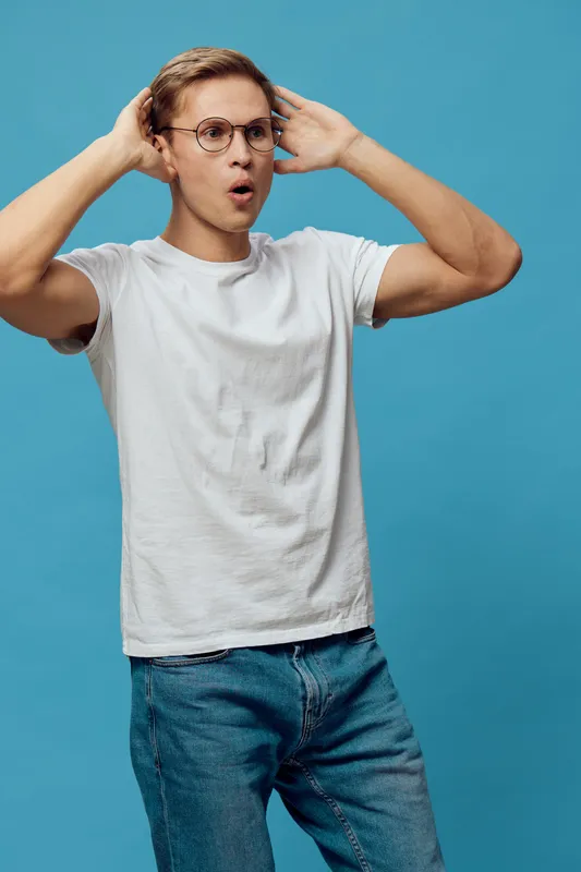 Confused young man with glasses wearing white t-shirt and blue jeans standing against blue background. Expression of surprise and uncertainty. Casual style, people emotion concept, modern fashion.