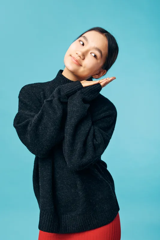 Young woman in black sweater standing with hands together near face smiling gently posing against blue background in studio portrait closeup with relaxed expression.