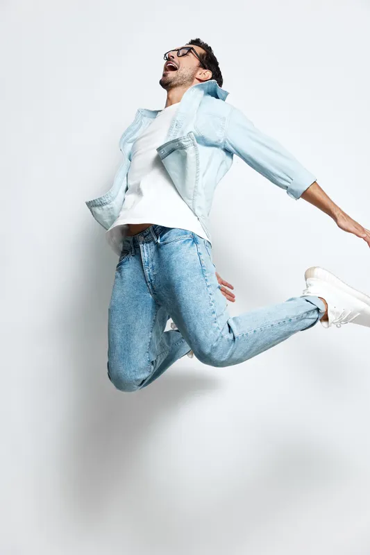 Energetic man jumping joyfully in casual outfit, wearing glasses and smiling, captured against a plain light background. Lifestyle concept of happiness and freedom.