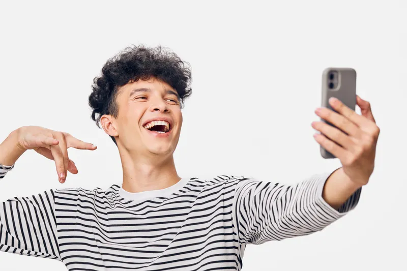 Young man taking a selfie with smartphone, laughing joyfully wearing striped shirt, isolated on white background, cheerful mood, casual style.