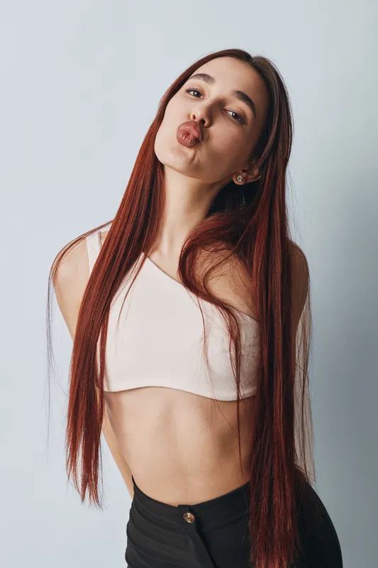 Young woman with long straight hair wearing white crop top and black pants posing against light background sending kiss with puckered lips and playful expression for lifestyle portrait.