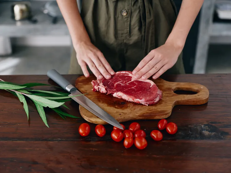 Женщина с ножом в руке нарезает свежее мясо для стейка для запекания на кухне, на столе соль, перец и другие специи, красные помидоры черри и зелень, приготовление ужина