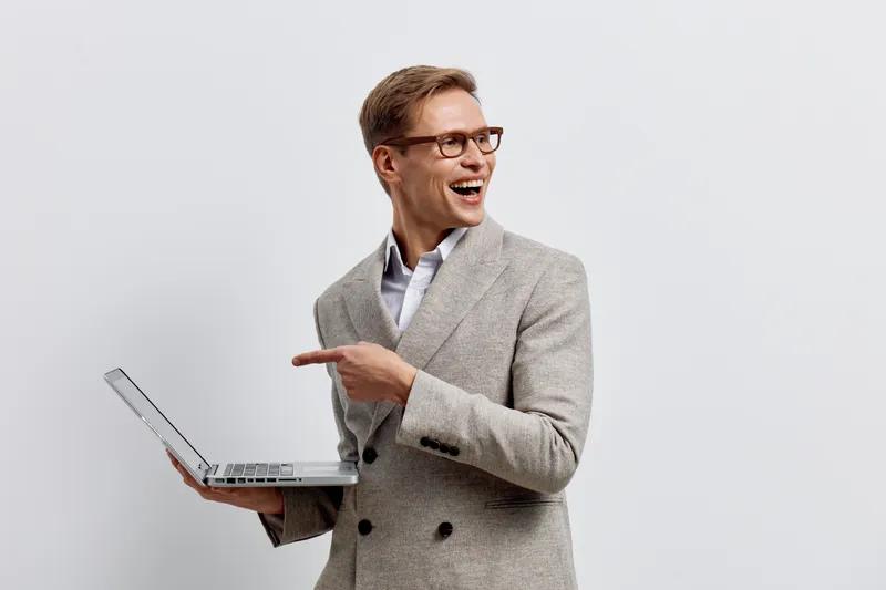 Confident smiling man in stylish gray blazer holding laptop, pointing to side, on plain white background. Business casual appearance, emotional expression, professional and positive atmosphere, studio