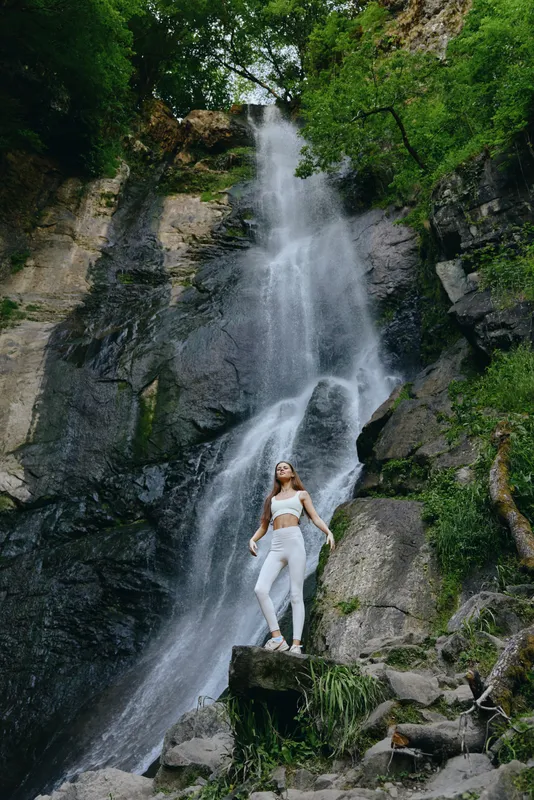 woman standing by tall waterfall in nature, wearing white activewear, surrounded by rocks and lush green trees, outdoor adventure and fitness concept