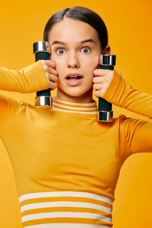 Woman fitness enthusiast holds dumbbells at shoulders against a vibrant orange background, wearing a striped top. Energetic workout scene highlighting strength, balance and focus during training.