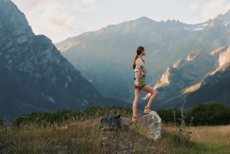 Woman hiking outdoors, standing on a rock in a sunlit valley, embracing nature, casual outfit, confident pose, scenic mountains, peaceful atmosphere