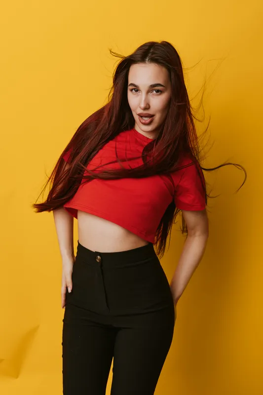 Young woman with long hair wearing red crop top and black pants posing confidently against yellow background, fashion style, casual outfit, studio portrait with dynamic hair movement.