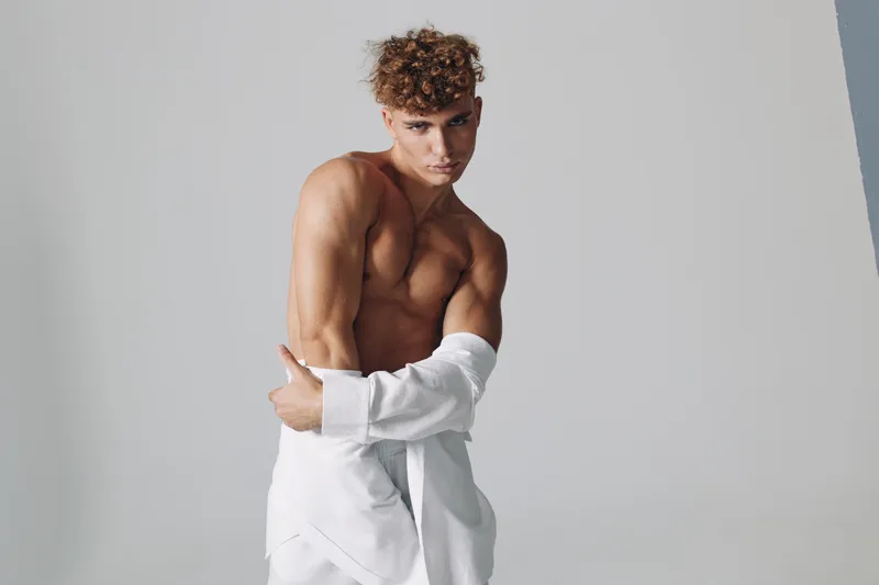 Muscular young man with curly hair removing white shirt posing in minimalist studio, showing athletic body and well-defined muscles, front and side view in bright environment.