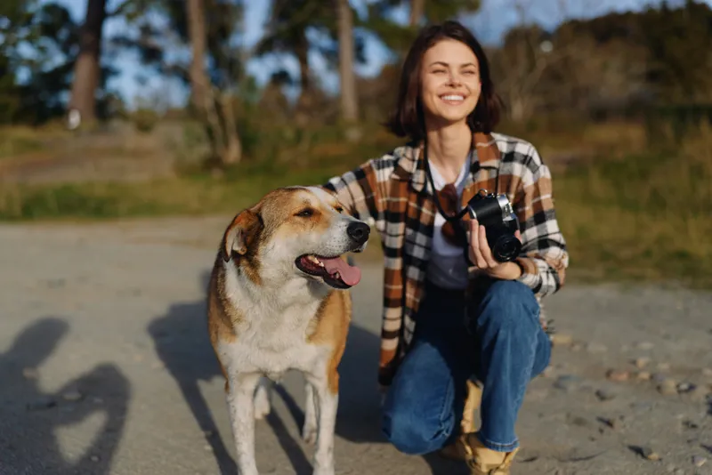 Woman kneels beside a friendly dog with a camera on a sunny outdoor trail, capturing candid moments and enjoying a peaceful park scene with soft light and warm companionship