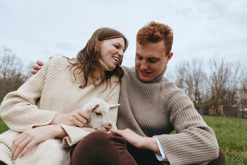 A smiling couple in cozy sweaters cuddle a baby goat in a sunny field, capturing a warm lifestyle moment of bonding, care, and rural living, with laughter and peaceful togetherness.