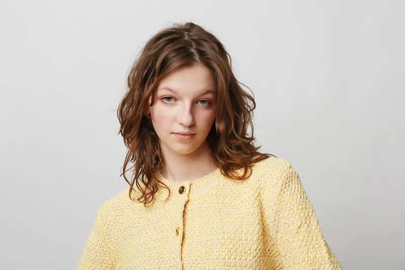 Young woman with curly brown hair wearing a yellow textured sweater looks confidently at the camera against a plain light background. Casual style and natural beauty concept.