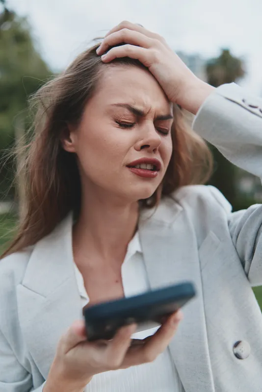 Business woman expressing frustration while holding a smartphone outdoors in a light-colored suit with a blurred background of greenery and soft cloudiness.