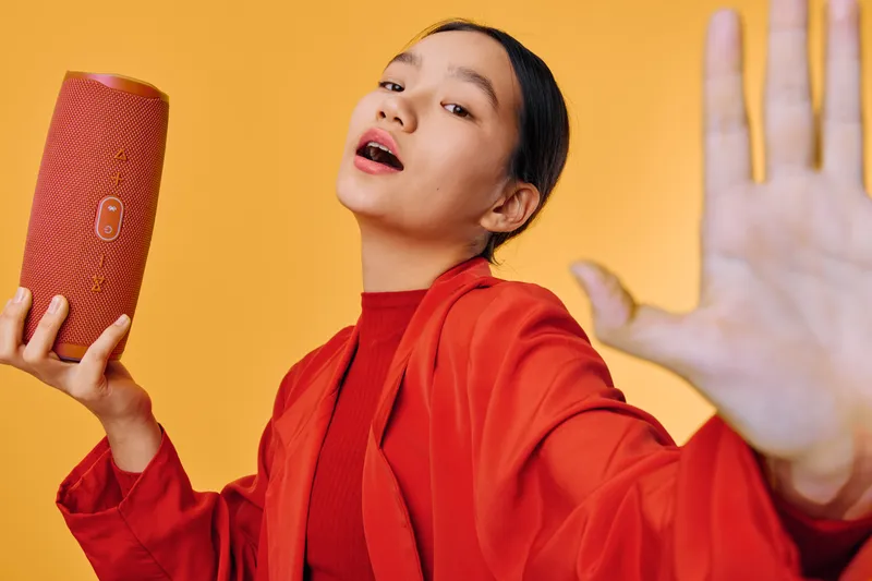Woman in red jacket holding portable speaker while extending hand against yellow background. Girl enjoying music with wireless sound device in casual pose, entertainment and technology concept.