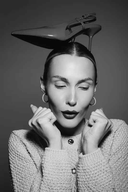 Fashionable white woman with light skin and dark hair holds a high heel shoe on her head, eyes closed, wearing knit sweater and hoop earrings in black and white studio portrait.