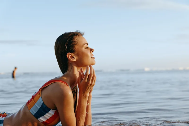 Young woman in striped swimsuit sits on the beach by the sea, hands near face, breathing calmly at sunrise. Ocean waves and morning sunlight show relaxed fitness outdoors.