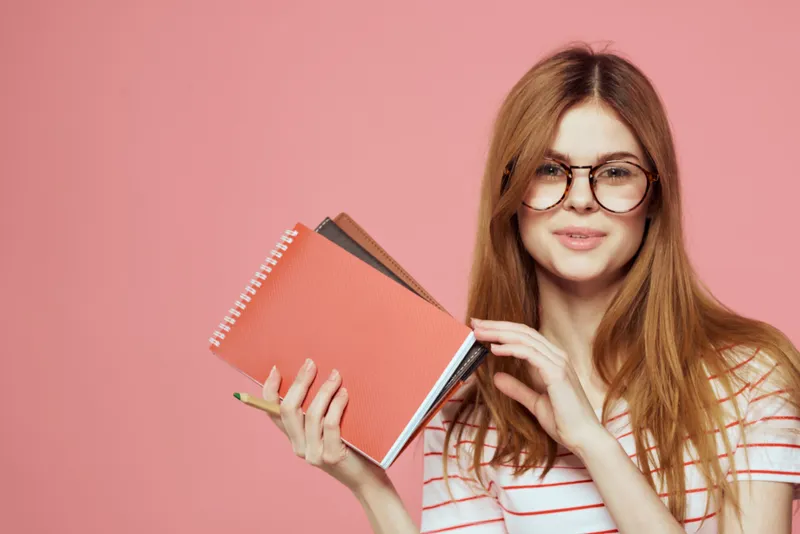 Красивая студентка держит книги, жестикулирует руками на розовом фоне