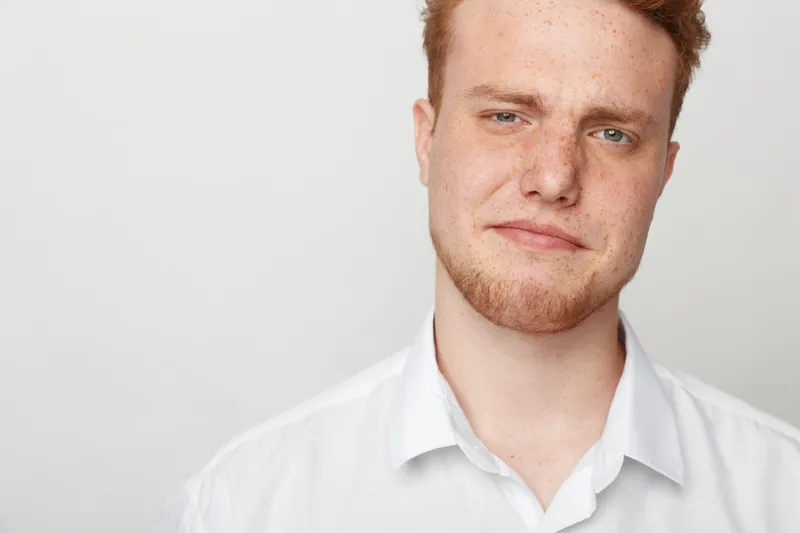Close-up portrait of a young redhead man with freckles and beard wearing a white shirt against a plain light background. Concept of natural beauty and casual style.