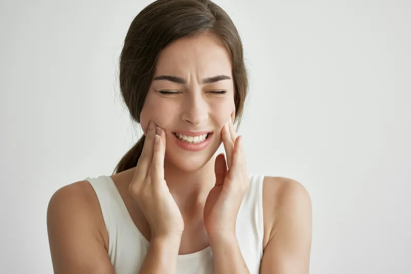 Женщина в белой футболке держится за лицо, испытывая зубную боль и недовольство, крупный план