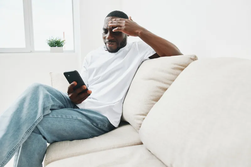 Stressed young African American man in casual white shirt checking phone on couch, showing emotions of anxiety and frustration
