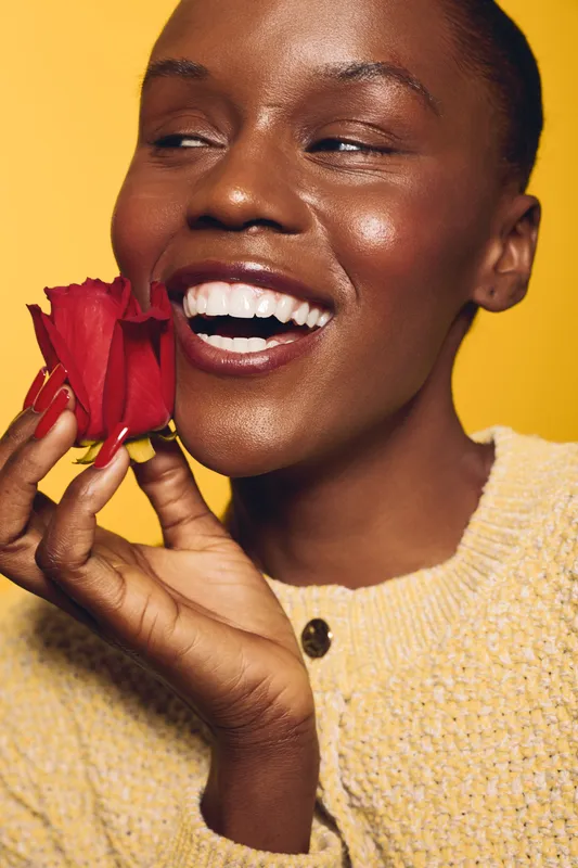 Smiling dark skinned African woman holds red rose near face, wearing yellow sweater. Close-up on yellow background. Concept of natural beauty, happiness, and floral skincare glow.