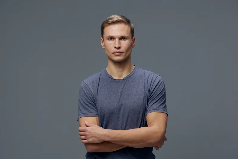 Confident young man with short hair wearing casual grey T-shirt looking directly at camera with crossed arms, standing against neutral grey background in studio portrait. People lifestyle concept