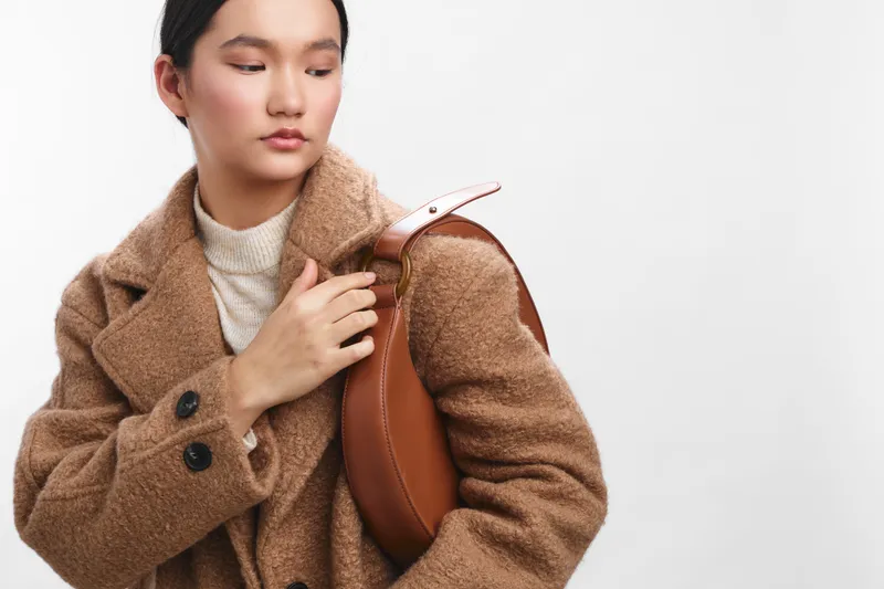 asian woman wearing a brown wool coat stands with a leather bag, looking down thoughtfully in a bright studio setting, conveying calm style and focused presence.