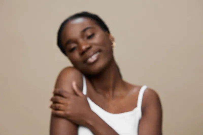 Young African woman smiling gently in a white tank top, embracing herself against a neutral beige background, expressing self love and confidence