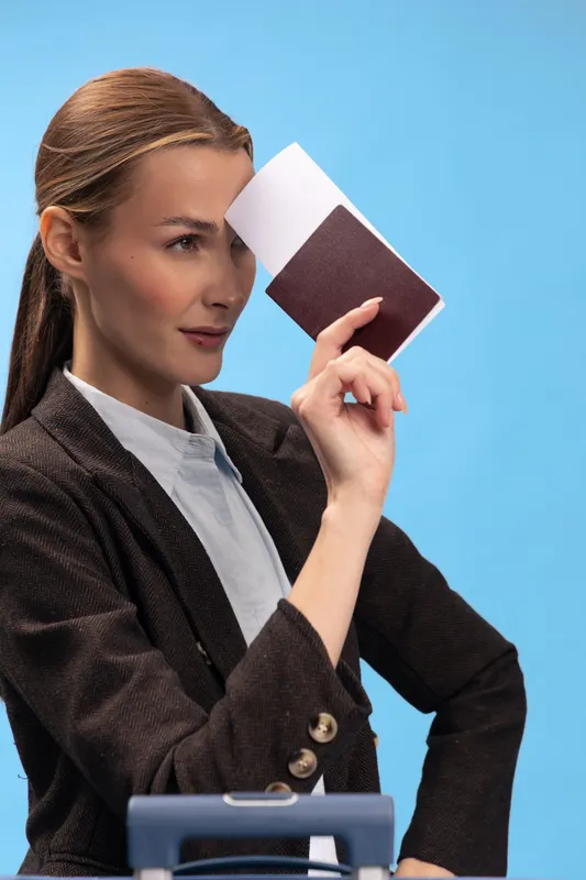 Confident woman in dark jacket holds passport near face with serious expression against blue background. Concept of travel preparation, identity verification, and business trip planning.