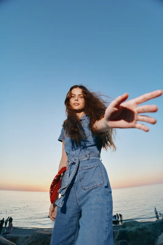 modern boho-western denim street style woman with long hair reaching out against sea and clear sky, film color, lifestyle fashion in natural light, casual summer vibe