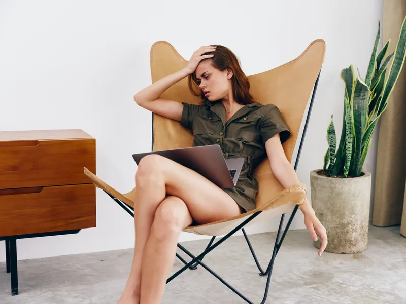 Женщина-фрилансер сидит в кресле с ноутбуком, работая из дома, испытывая грусть, недовольство и усталость