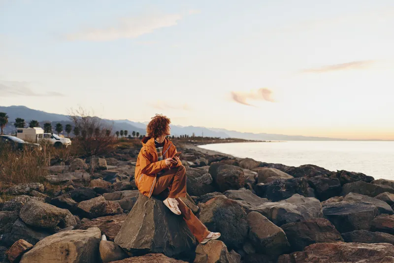 Woman lifestyle by the seaside, sitting on rocks at sunset. Curly hair in an orange jacket, relaxed posture and outdoor travel mood. Golden sky, soft calm ocean water, warm distant hills.