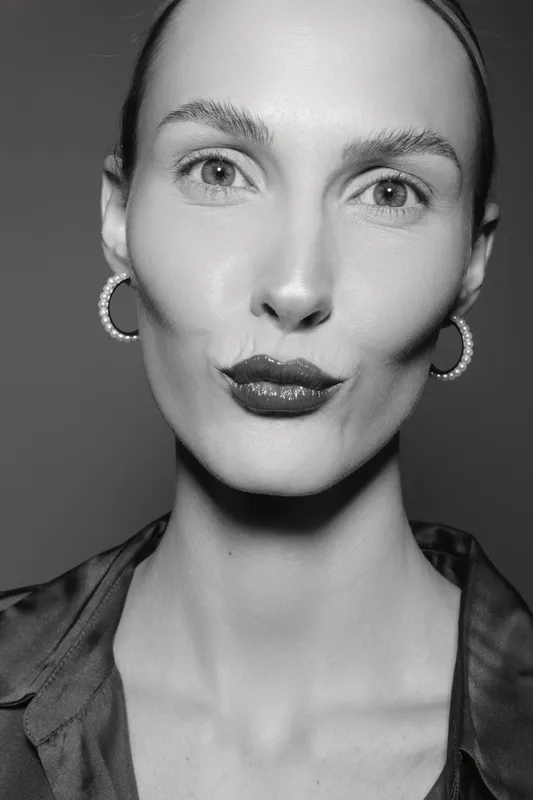 Close-up portrait of a pale white woman with sharp facial features and slicked-back hair wearing pearl earrings and dark shirt. Studio monochrome beauty concept.