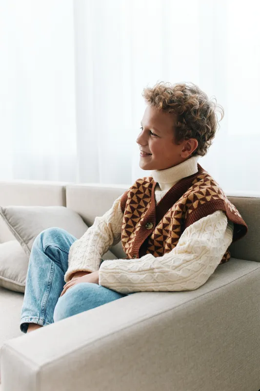 A relaxed person sits on a light sofa wearing a cream turtleneck sweater and a patterned knit vest, smiling softly as soft natural light streams through a bright window.