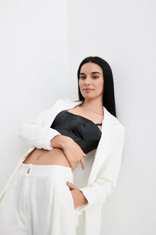 Young woman wearing white suit and black top poses confidently with long black hair against a white minimalist background in a modern studio setting.