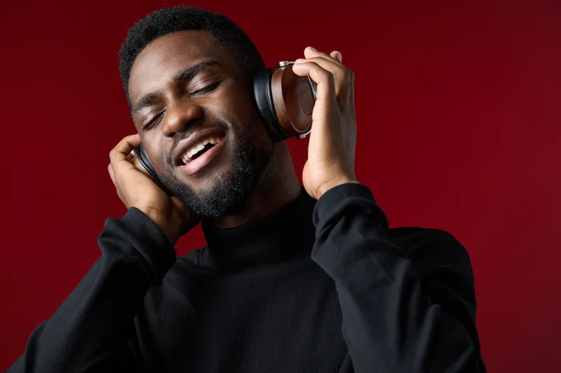 Young Black man enjoying music with headphones against a rich red background, expressing joy and bliss while immersed in sound