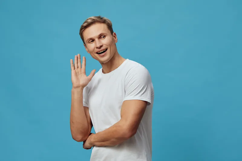 Smiling young Caucasian man with short light brown hair wearing plain white t-shirt, waving hand and looking cheerful against a bright blue background in a studio portrait. People lifestyle concept, h
